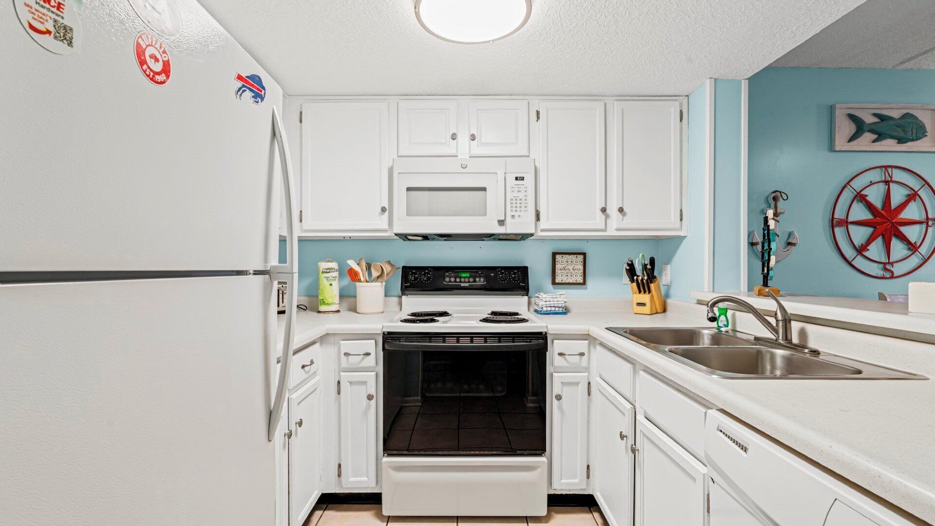 Une cuisine avec des meubles blancs, un four à micro-ondes, une cuisinière et un évier. On y trouve des objets décoratifs sur le mur, ainsi qu’une décoration murale sur le thème de la marine.