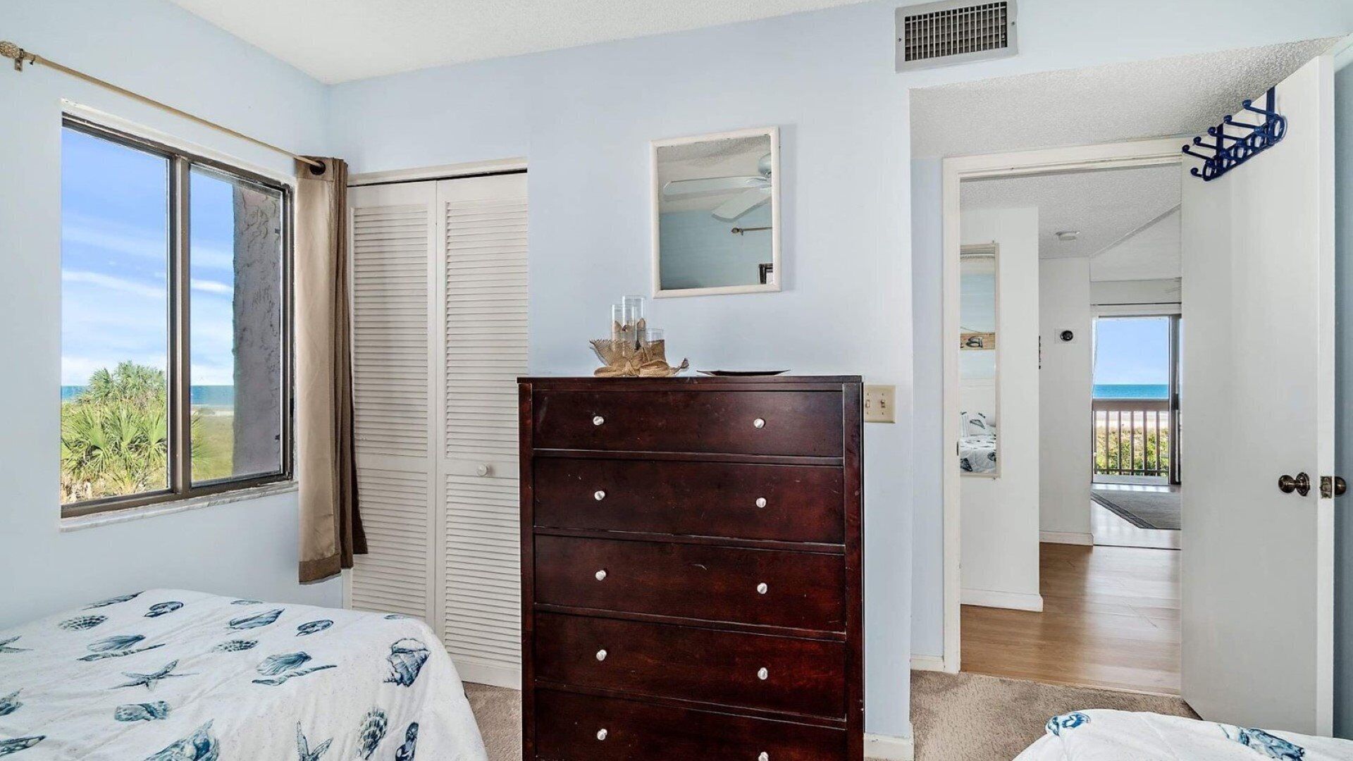 A bedroom with a bed, a dresser, a window with a view, a door leading to a balcony, and a small mirror on the wall.