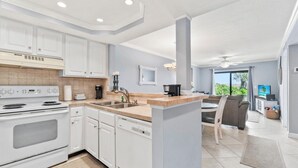 A modern kitchen with white cabinets, a built-in oven, and a sink. There is a dining area with a table and chairs, and a large window offering a view of greenery. - Beachfront Community Condo – Dune View, Easy Beach Access, Resort Amenities (St. Augustine)