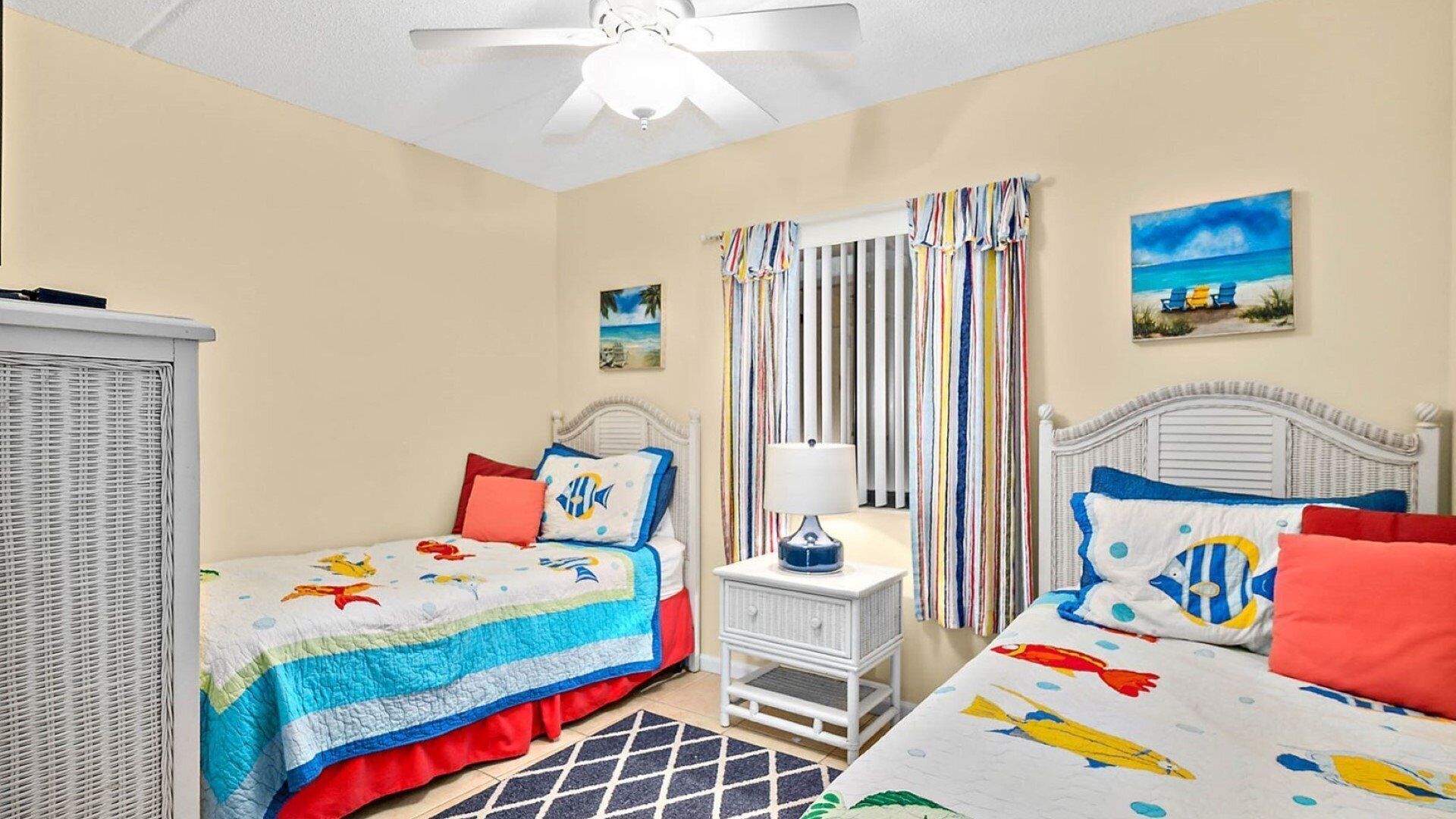 A bedroom with two beds, striped curtains, a ceiling fan, and a beach-themed painting.
