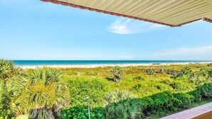 Uitzicht vanaf een balkon op een strand, met een houten hek en weelderige begroeiing.