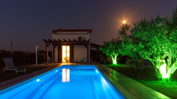 Una piscina con pérgola y sillas de descanso, rodeada de follaje e iluminada con luces verdes.