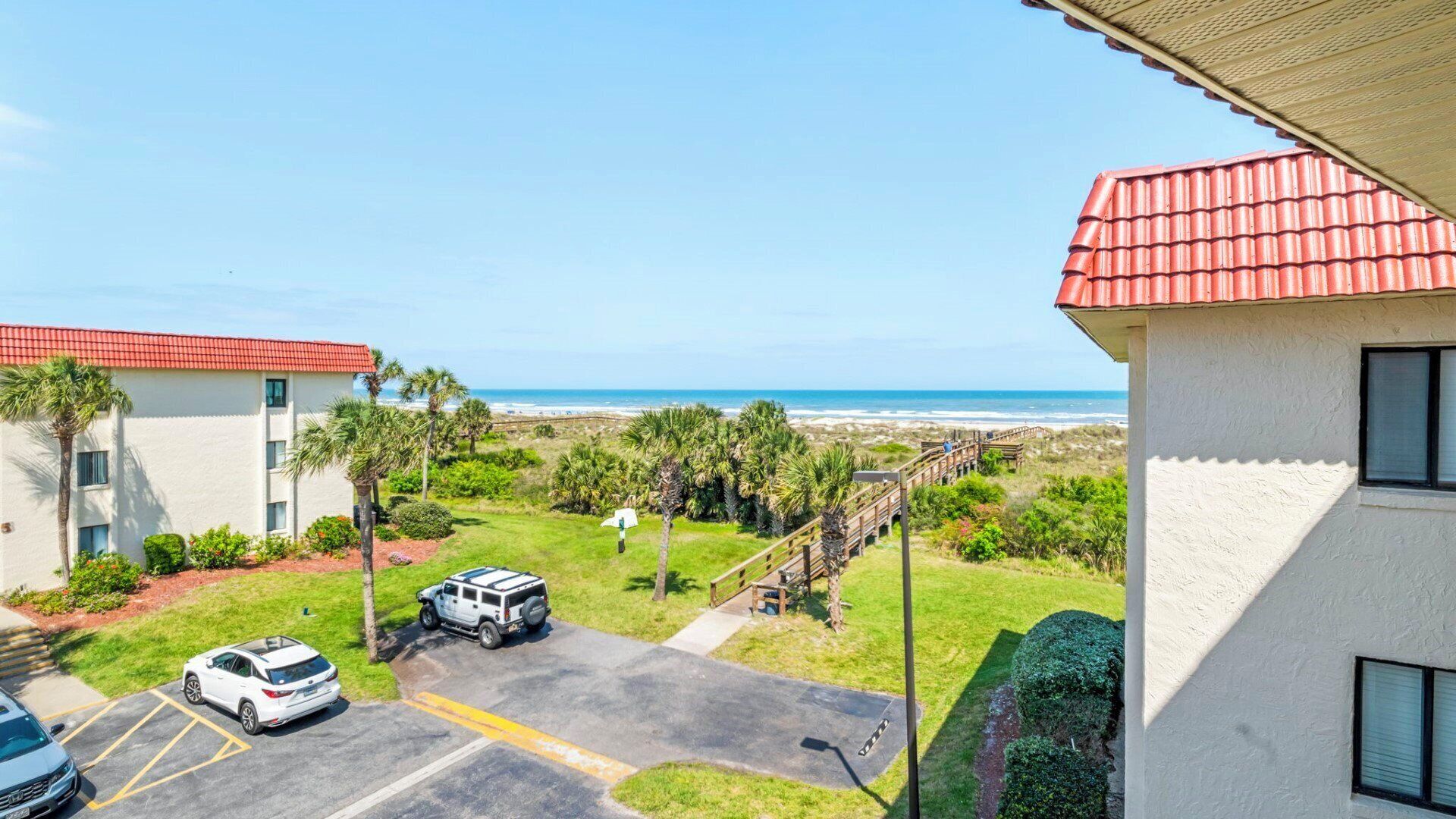 A hotel with a red-tiled roof, a parking area with two cars, and a view of the beach and ocean.