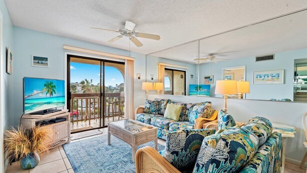 A living room with a large TV, a sofa, a coffee table, and a balcony with a view. - Beautiful Ocean-View St. Augustine Condo • Steps from Beach (St. Augustine)
