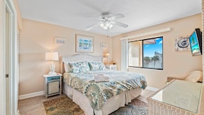 A bedroom with a bed, a ceiling fan, a window with a view, and a TV on the wall.