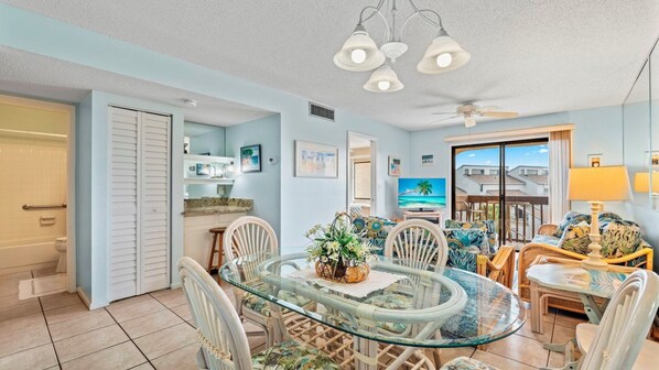 A spacious living area with a glass dining table surrounded by wicker chairs, a comfortable sofa with tropical cushions, and a ceiling fan.