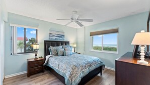Un dormitorio con cama, dos mesitas de noche, ventilador de techo y ventanas amplias que ofrecen vistas al exterior.