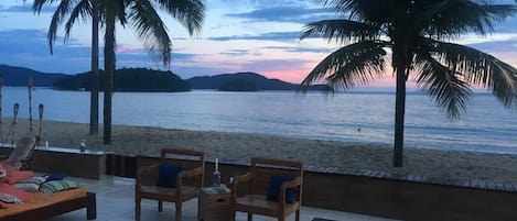 A beachside patio with wooden furniture, a bench, and a view of the sea and distant hills.