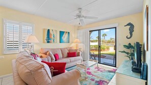 A living room with a sofa, a coffee table, and a TV stand. There are two lamps, a ceiling fan, and a large window with blinds. - Poolside Condo – Steps to Beach, Free Activities, Near Historic St. Augustine (St. Augustine)