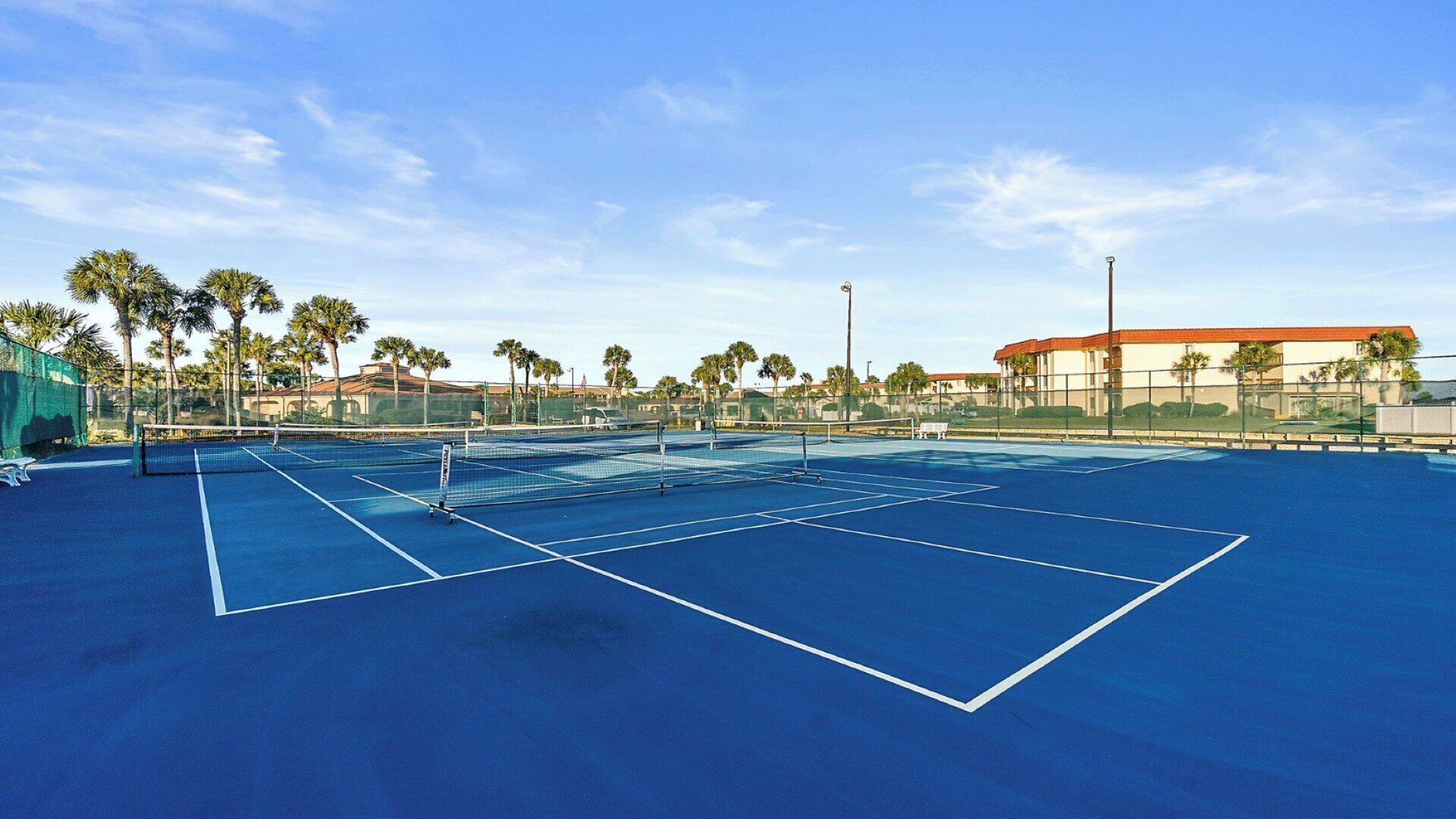 An outdoor tennis court with a blue surface, surrounded by palm trees and buildings.