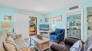 A living room with a light blue wall, a television, a blue armchair, and a white door labeled 'ST. AUGUSTINE.' - Poolside 2BR Condo in Oceanfront Resort - Sleeps 6, Steps to Beach (St. Augustine)