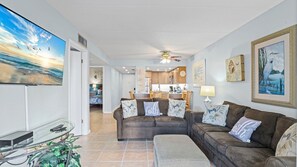 A living room with a sofa set, a coffee table, and a TV screen displaying a beach scene. There is a kitchen in the background with wooden cabinets and a dining area. - 5115 Ocean Side 1st Floor NE (St. Augustine)