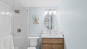 Une salle de bain avec des toilettes blanches, une douche à pommeau large, un lavabo en marbre avec un miroir et un meuble en bois.