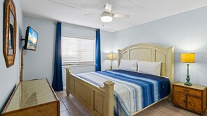 Une chambre à coucher comprenant un lit, des lampes de chevet, un ventilateur de plafond, une fenêtre avec des stores et un miroir.