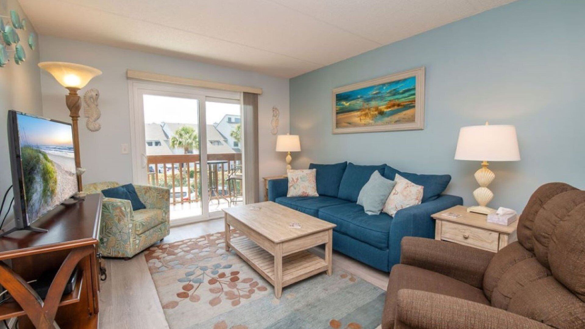 A living room with a blue sofa, a wooden coffee table, a patterned armchair, and a television on a wooden stand. There is a painting on the wall and a window with a view of buildings.