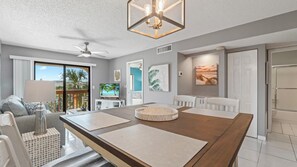 A spacious living area with a wooden dining table, a ceiling fan, and a large window offering a view of palm trees.