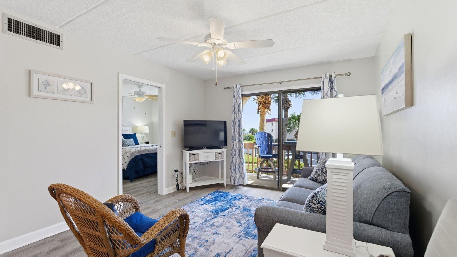 A spacious living area with a ceiling fan, a TV on a white stand, a grey sofa, a wicker chair, and a blue patterned rug.