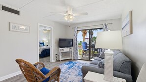 A spacious living area with a ceiling fan, a TV on a white stand, a grey sofa, a wicker chair, and a blue patterned rug.
