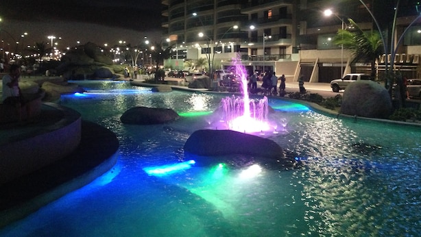 Una piscina nocturna con fuentes iluminadas y edificios circundantes.
