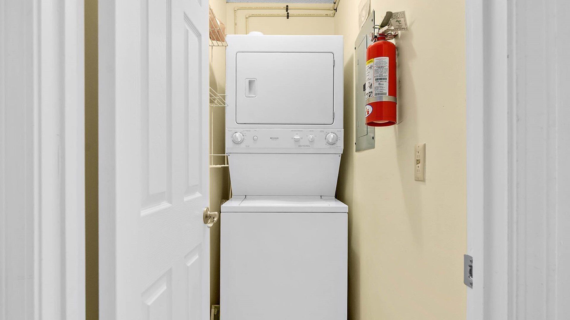 A laundry room with a white washer and dryer, a fire extinguisher mounted on the wall, and a door leading to another room.