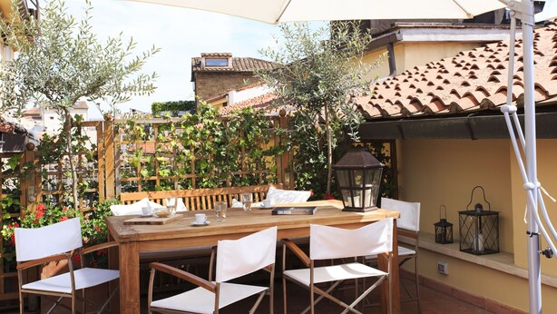 Un patio exterior con una mesa de madera y sillas blancas, rodeado por un cerco de madera y follaje.