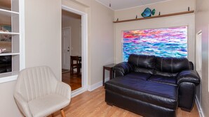 A living room with a black leather sofa, a white armchair, and a colorful abstract painting on the wall. - Seabird Shack: Dog Friendly, Downtown Pensacola! (Pensacola)