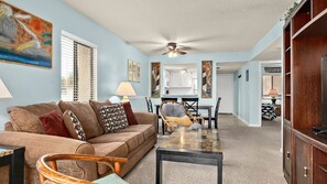 A living room with a sofa, coffee table, and chairs. There is a dining area with a table and chairs. The room has a ceiling fan and artwork on the walls.