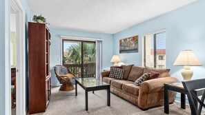 A living room with a sofa, two armchairs, a coffee table, and a bookshelf. There is a window with blinds, a painting on the wall, and a lamp on a side table.