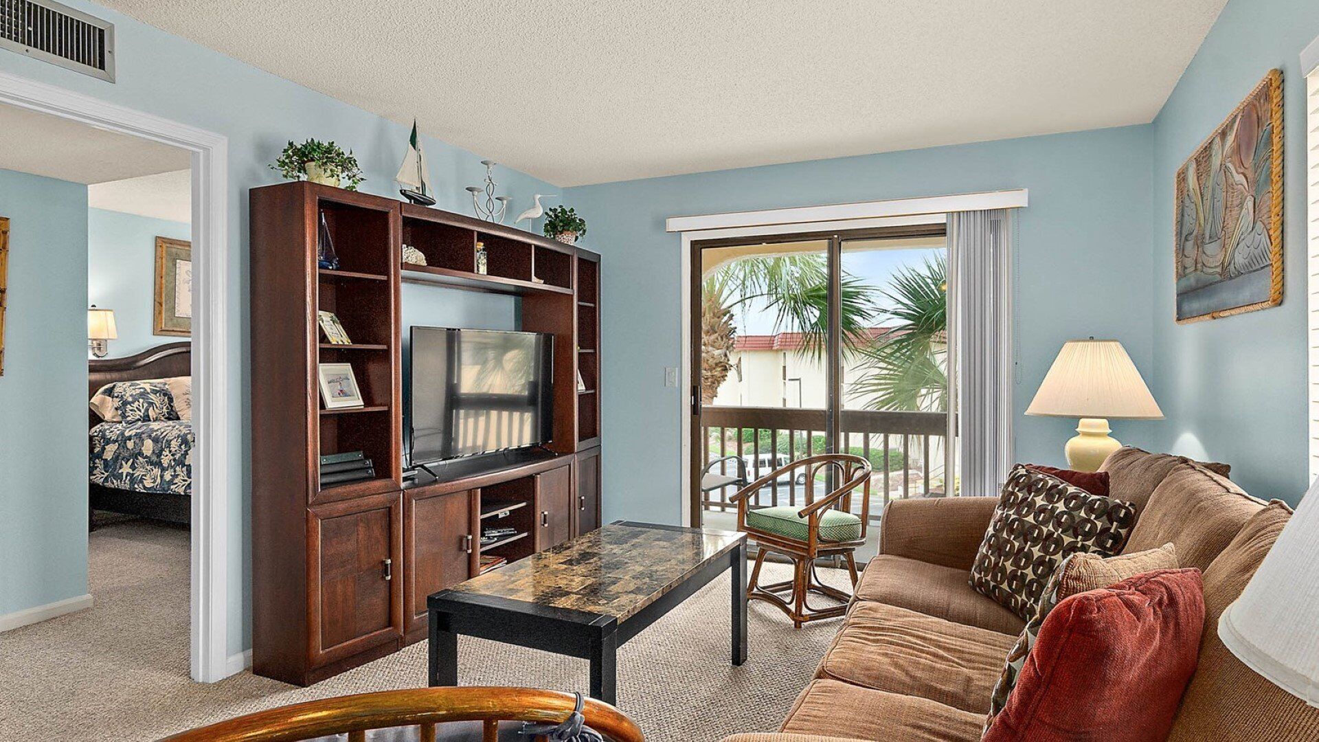 A living room with a TV, wooden entertainment center, and a balcony with a view of palm trees.