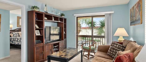 A living room with a TV, wooden entertainment center, and a balcony with a view of palm trees.