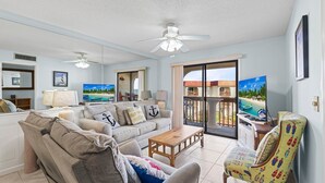 Uma sala de estar espaçosa com um sofá grande, uma mesa de centro e uma poltrona. Há uma varanda com vista para a praia e um edifício.