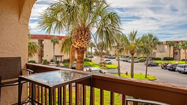 A balcony with a glass table and chairs, overlooking a parking area with palm trees and a building with a red roof.