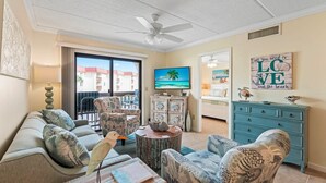 Een woonkamer met een bank, fauteuils, een salontafel, een televisie en een balkon met uitzicht op gebouwen.
