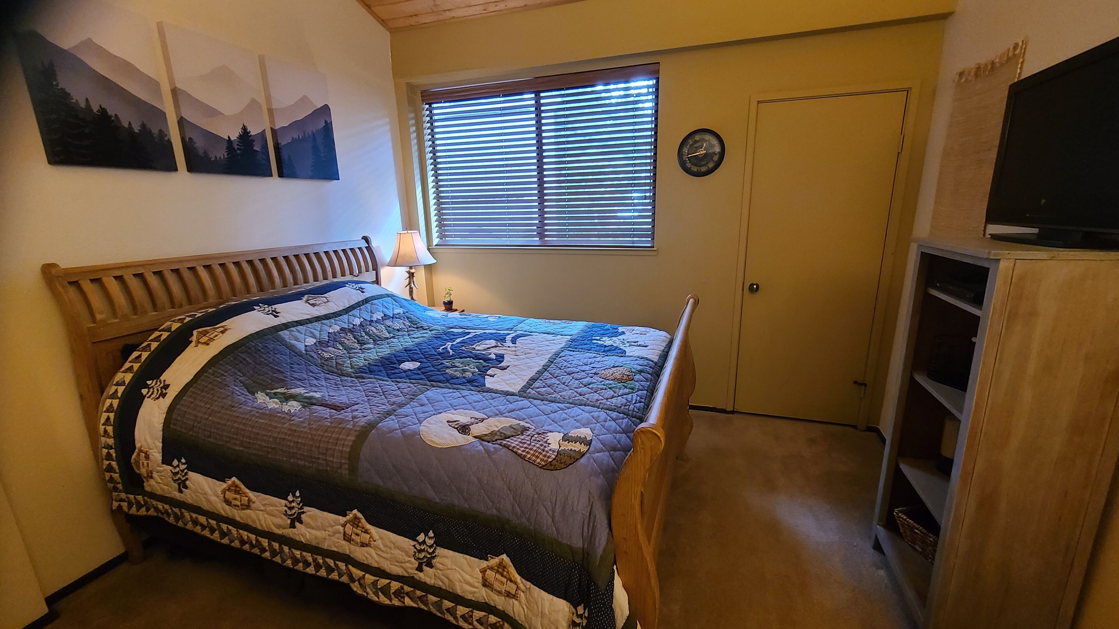 Et soveværelse med en stor seng, et træsengegavl, et vindue med rullegardiner, et ur på væggen og et fjernsyn på et træskab.