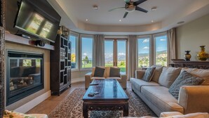 A spacious living room with a fireplace, a television, a ceiling fan, and large windows offering a scenic view. - Grand Vineyard Estate Home Exceptional Place Best Location in Penticton (Penticton)