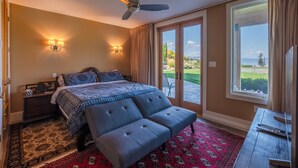 A bedroom with a bed, two wall-mounted lamps, a ceiling fan, a large window with curtains, and a view of the outdoors. - Grand Vineyard Estate Home Exceptional Place Best Location in Penticton (Penticton)