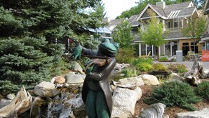 Eine Statue einer Person in einem grünen Anzug, die einen Bogen und einen Pfeil in einem Garten mit Teich, Steinen und Bäumen hält.