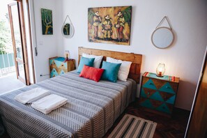 Une chambre à coucher bien aménagée, avec un grand lit, des coussins colorés, une tête de lit en bois, une table de chevet avec une lampe et des œuvres d’art accrochées aux murs.