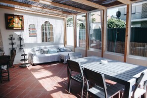 Une terrasse ensoleillée avec une table à manger, un coin salon confortable et une grande fenêtre donnant sur l’extérieur.