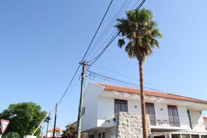 Un bâtiment à deux étages, doté d’une façade blanche et d’un toit recouvert de tuiles rouges. On y trouve un balcon avec une balustrade et une petite table.