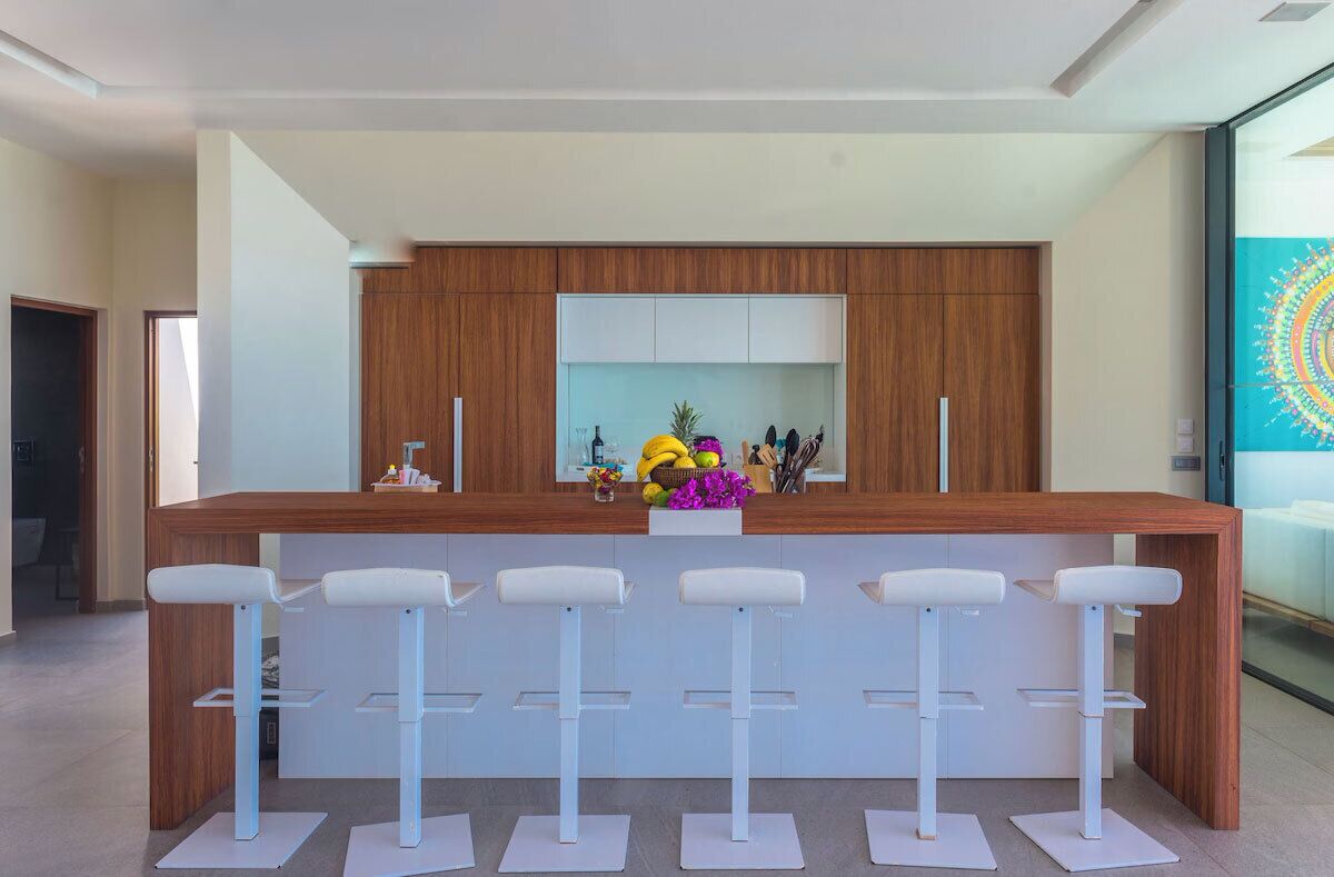 A modern kitchen with a wooden countertop, white bar stools, and a central island with a fruit bowl and decorative items.