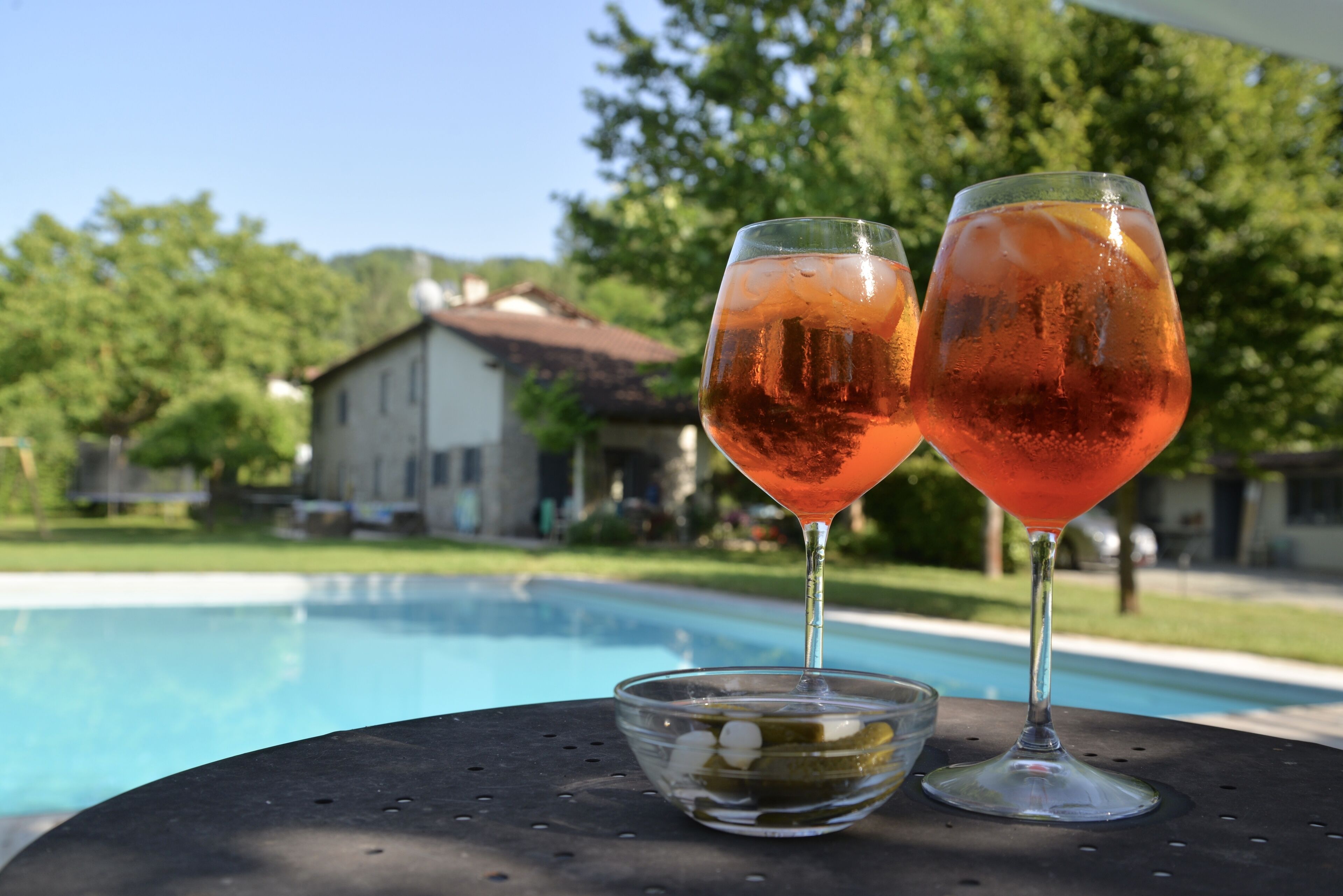 Two glasses of pink beverage on a table by a swimming pool with a house in the background.