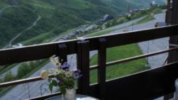 Ein Balkon mit Glastisch und Blumenvasen, mit Blick auf eine Berglandschaft und eine Straße.
