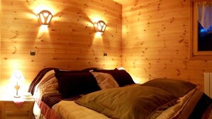 A wooden cabin room with a bed, two wall-mounted lights, a window with curtains, and a radiator. - Luxury chalet in the Savoie, typical of the Beaufortain area -classified 4 (Villard-sur-Doron)