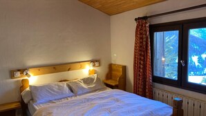 A bedroom with a wooden headboard, a bed with white bedding, a bedside table, a chair, and a window with curtains. - LARGE chalet with garden in Molines-en-Queyras, sleeps 19, fully equipped. (Molines-En-Queyras)