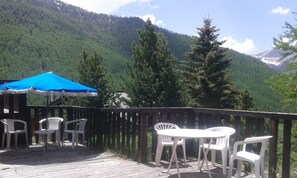 A wooden deck with white plastic chairs and a table, overlooking a forested mountain range. - LARGE chalet with garden in Molines-en-Queyras, sleeps 19, fully equipped. (Molines-En-Queyras)