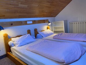 A room with two wooden beds, white bedding, a wooden headboard, a shelf unit, and a radiator. - LARGE chalet with garden in Molines-en-Queyras, sleeps 19, fully equipped. (Molines-En-Queyras)
