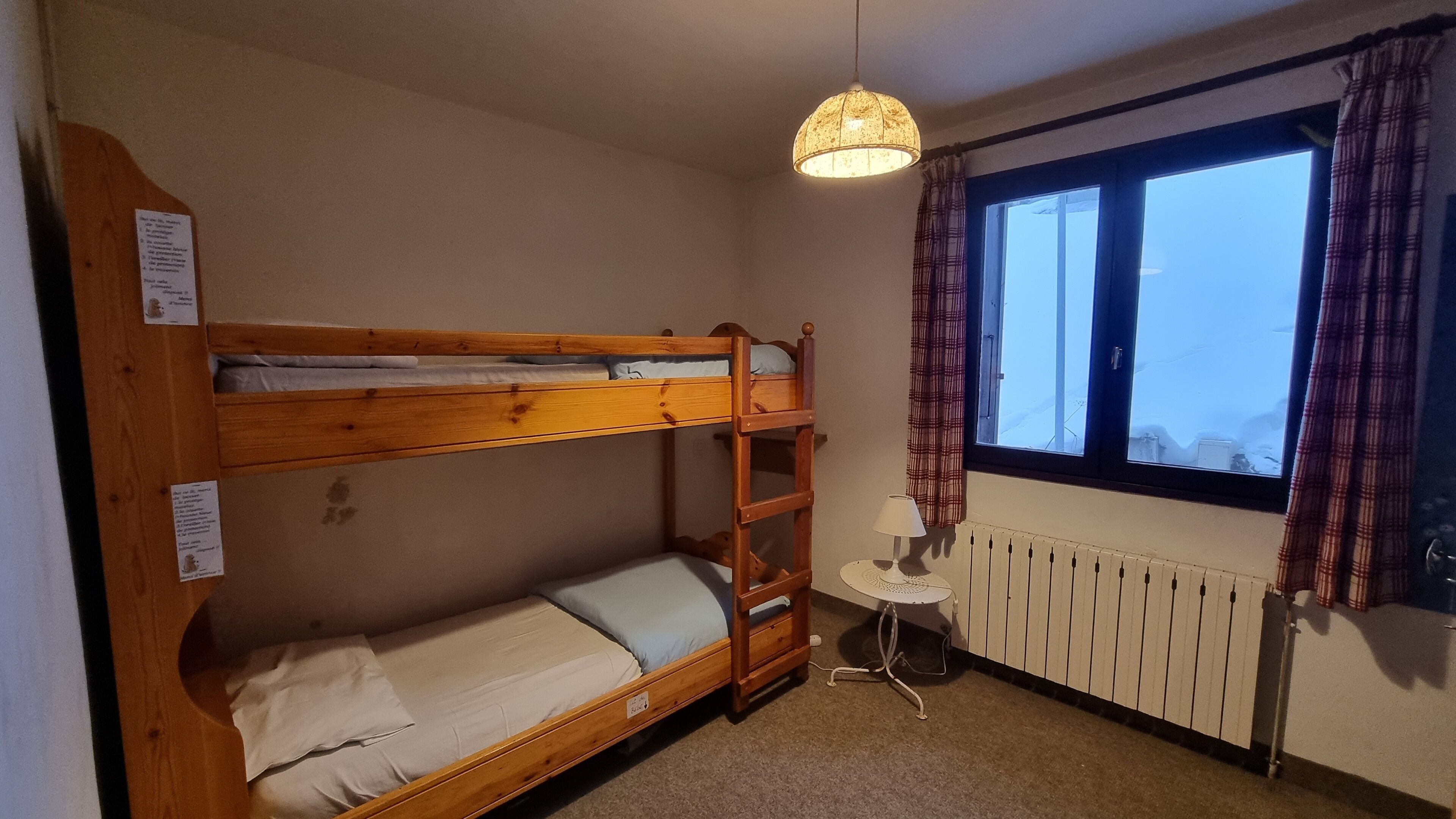 A bunk bed room with a radiator, a small table with a lamp, and a window with curtains.