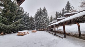 A snow-covered outdoor area with wooden picnic tables and benches, surrounded by tall evergreen trees. - LARGE chalet with garden in Molines-en-Queyras, sleeps 19, fully equipped. (Molines-En-Queyras)