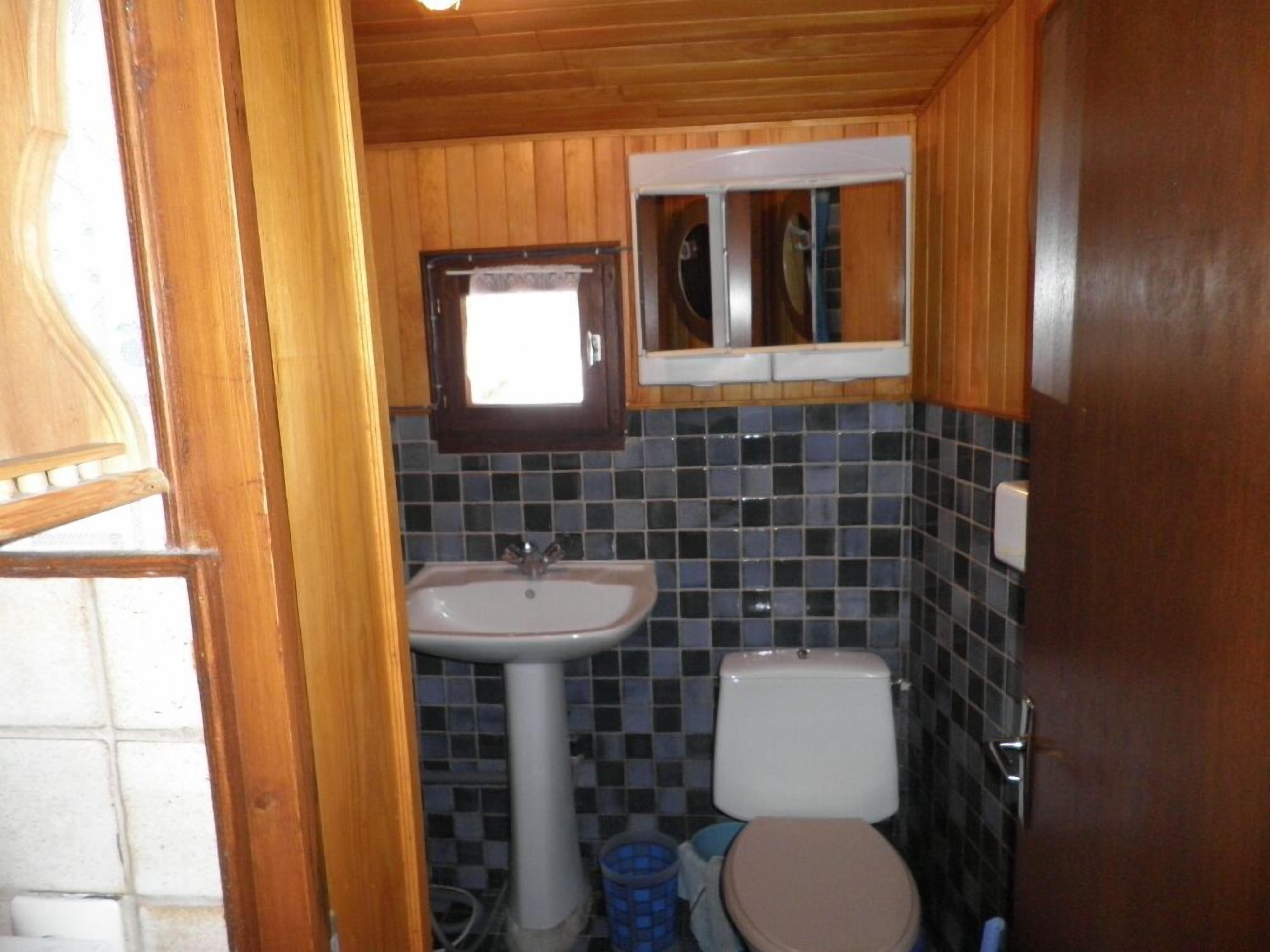 Un baño pequeño con revestimiento de madera, un lavamanos pedestal blanco, un inodoro y una ventana sobre el lavamanos.